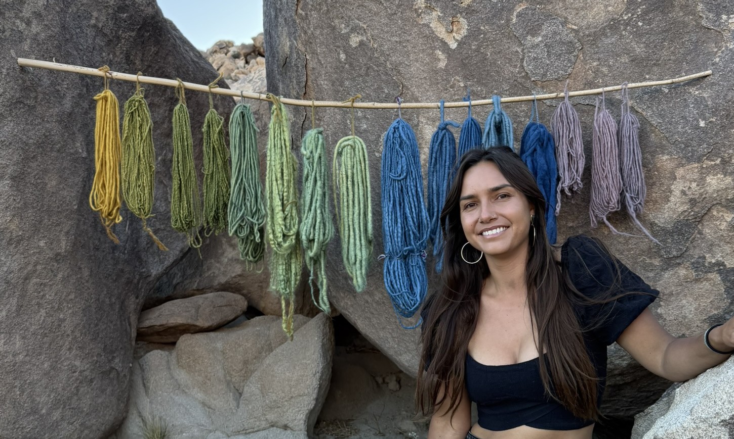 Plant-dyed yarn skeins in yellow, green, blue, and violet hanging from a pole against desert boulders