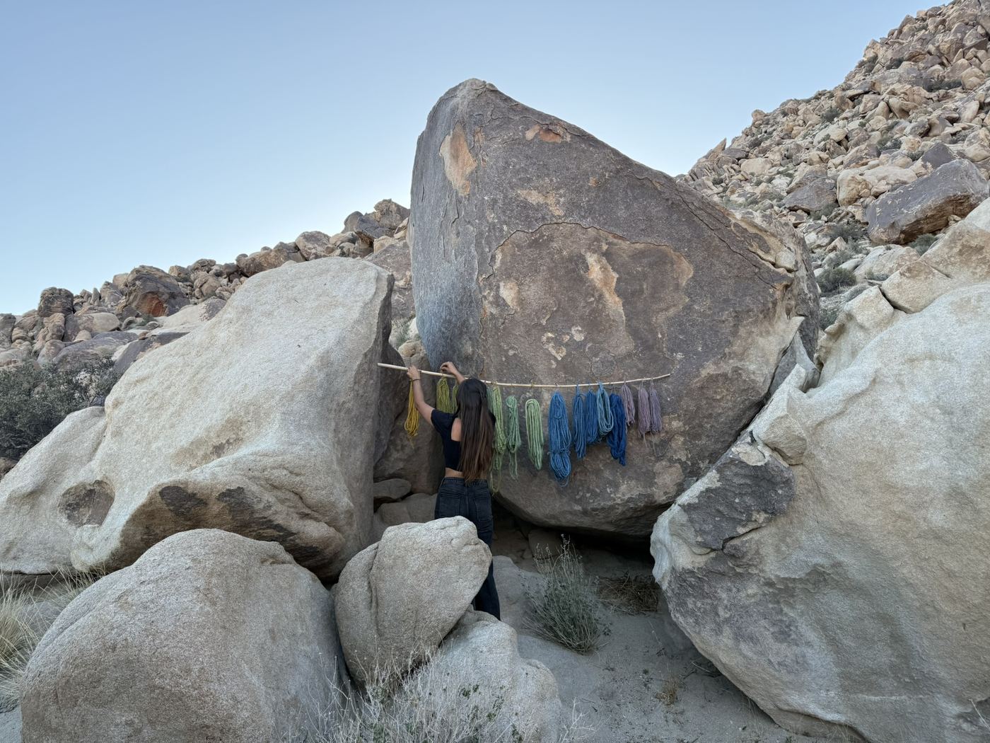Felise hanging a pole of plant-dyed yarn skeins against desert boulders