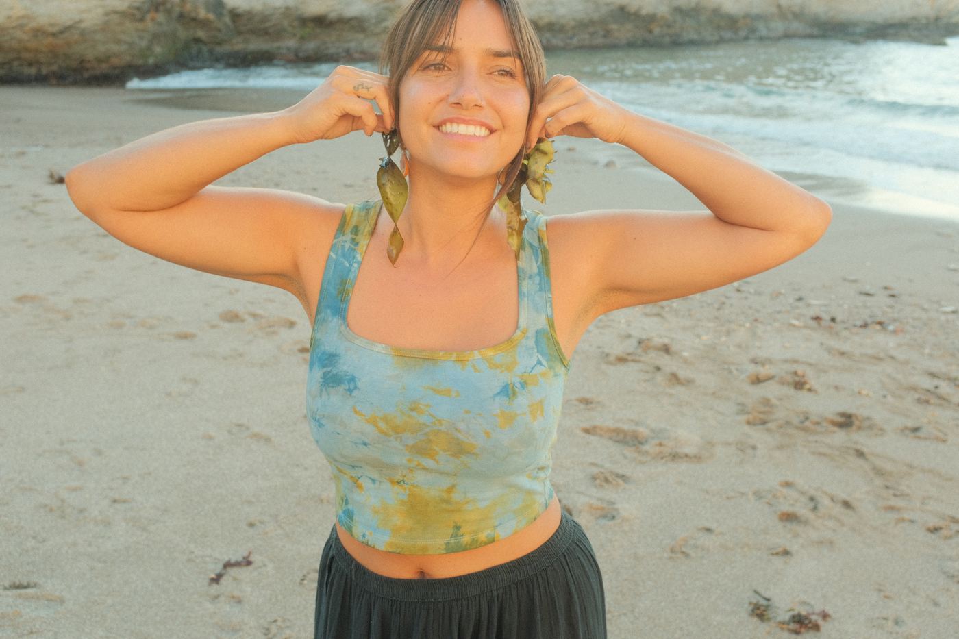 Felise on the beach in a blue-green plant-dyed tank, holding seaweed to her ears