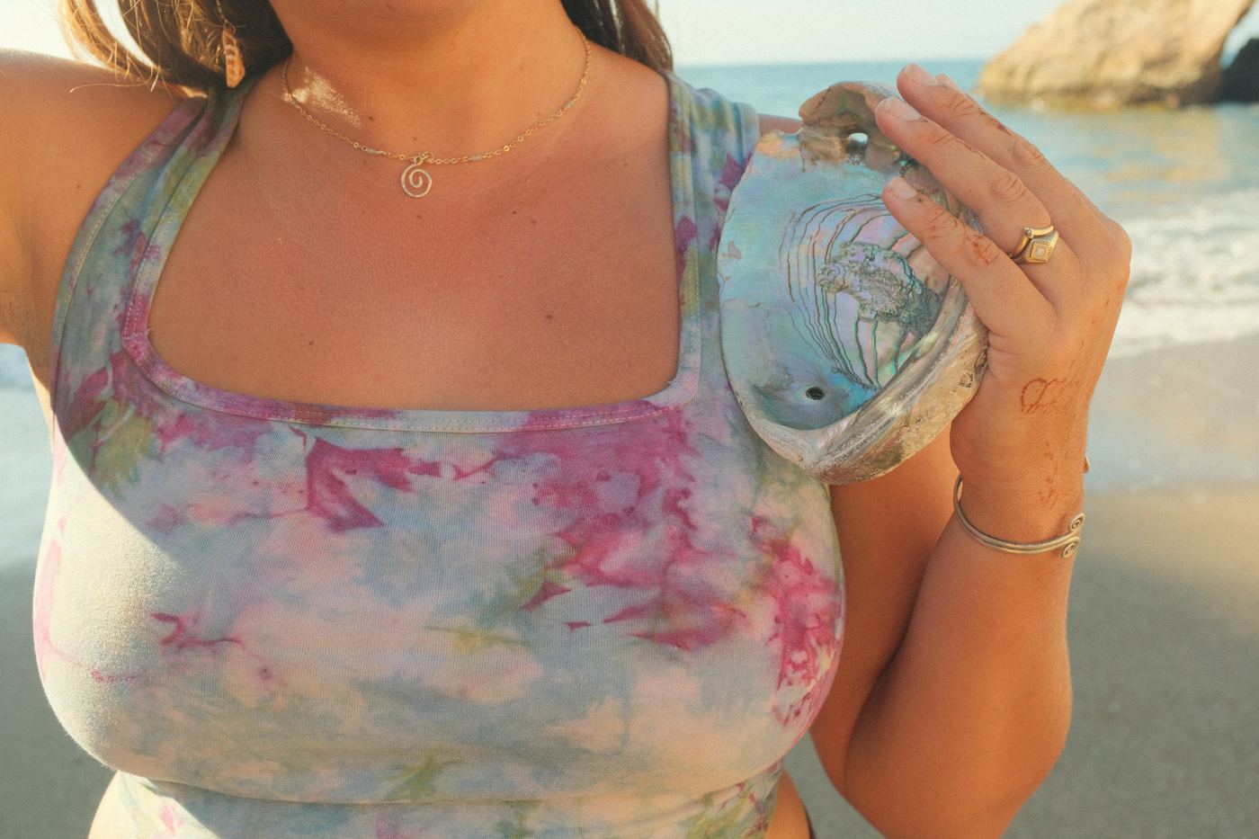 Close view of a pink plant-dyed tank, spiral necklace, and an abalone shell held at the shoulder