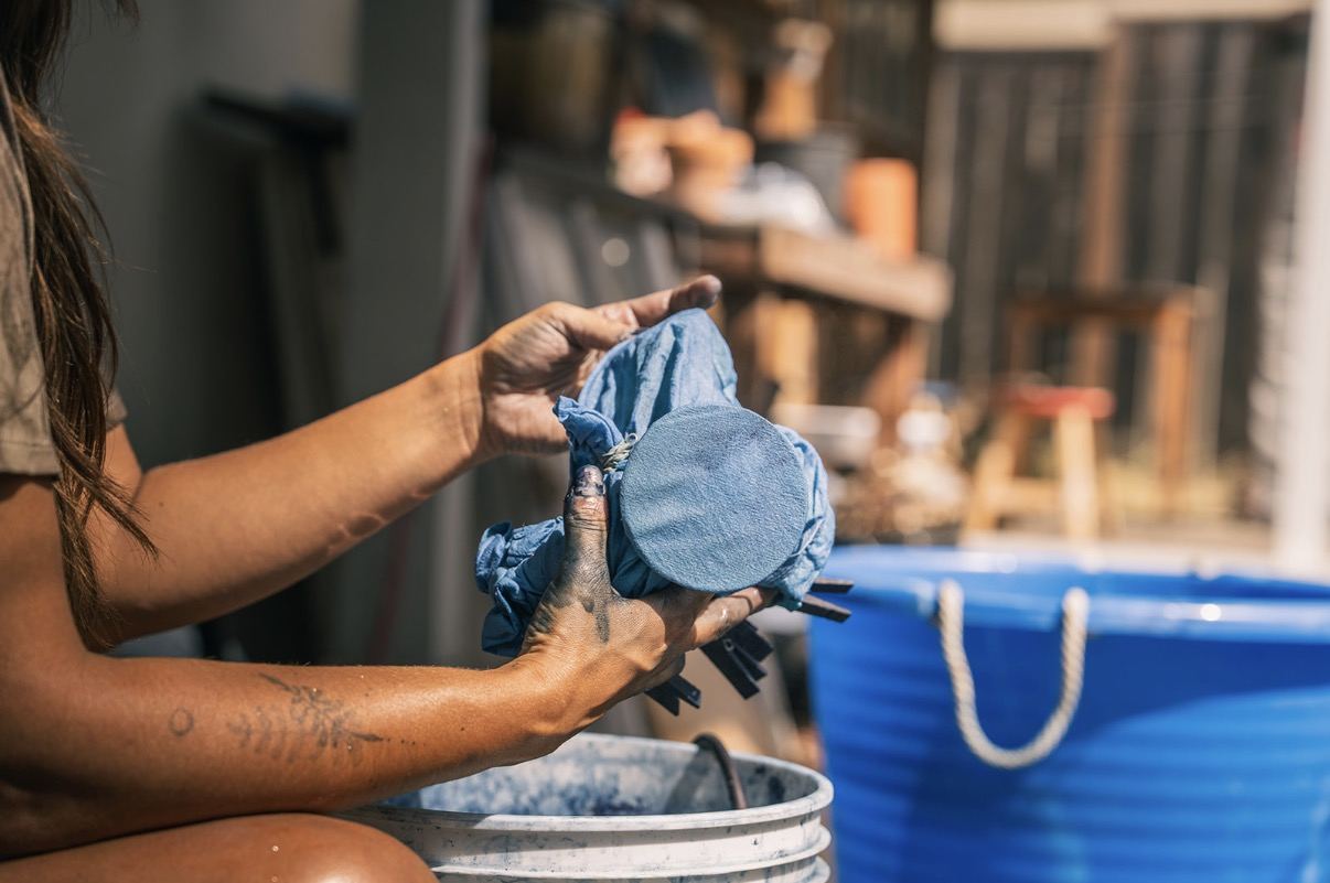 Indigo-stained hands wrapping cloth around a disc beside a dye bucket